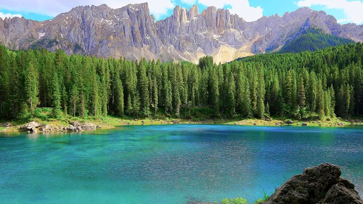 A stunning view of a lake surrounded by lush green trees and majestic mountains in the background, with a bright blue sky dotted with white clouds