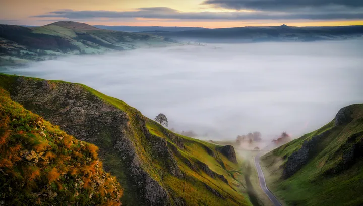 A misty sunrise over the Yorkshire Dales in the Peak District of England, with rolling hills covered in grass and a tree in the foreground, and a sky 