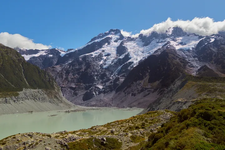A stunning view of a mountain range with a lake in the foreground, surrounded by lush green grass and snow-capped peaks