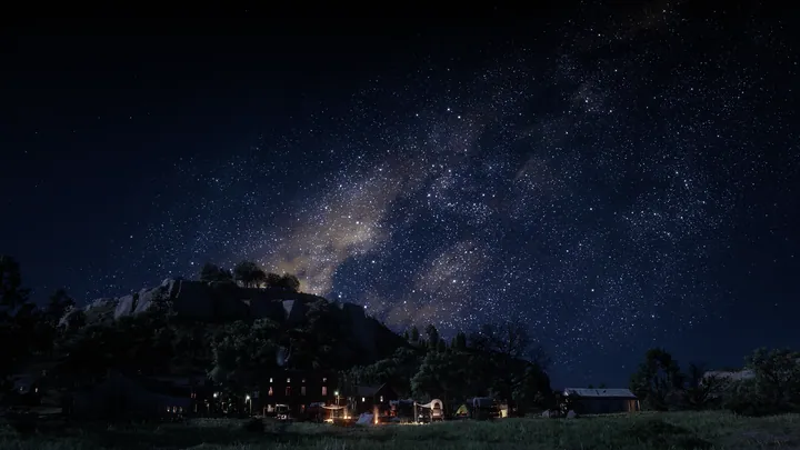 A night sky filled with stars, illuminated by the milky way, over a grassy field with houses and trees in the foreground and a mountain in the backgro