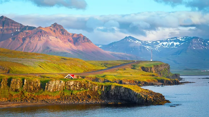 A small house perched atop a rocky cliff overlooking a body of water, with mountains in the background and a sky filled with clouds