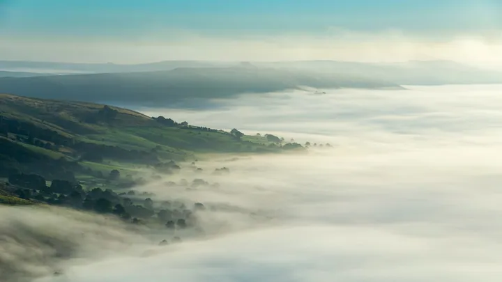 A view from the top of a hill above the clouds, with trees and hills visible below