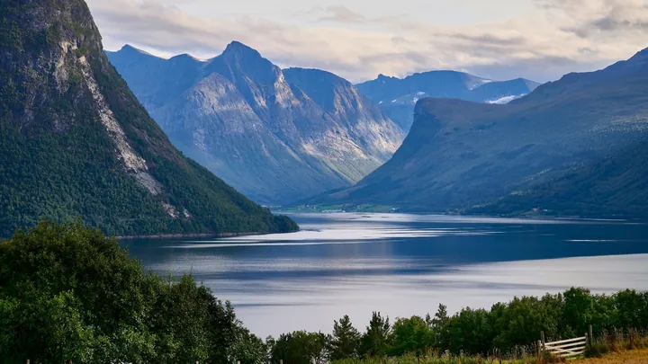 A large body of water surrounded by lush green trees and grass, with majestic mountains in the background and a sky filled with white, fluffy clouds
