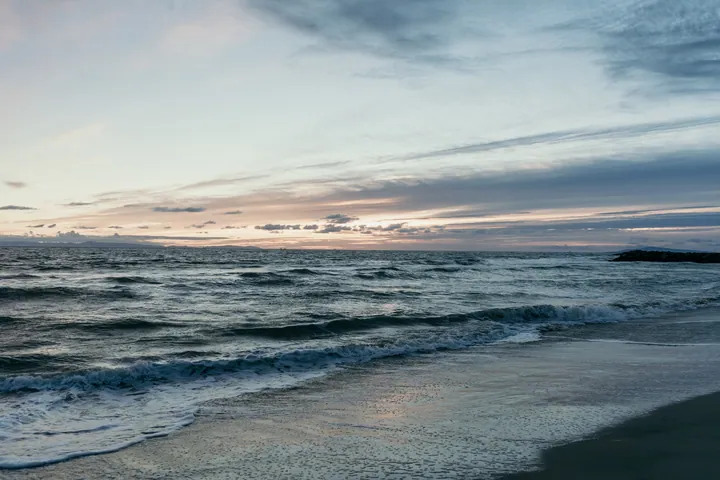 A beach with waves crashing onto the sand at sunset, with the sky in the background filled with clouds