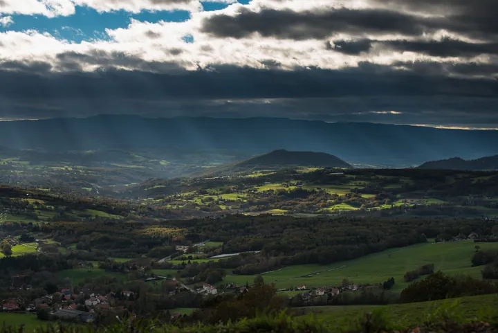 A view from the top of a hill with lush green grass, trees, and hills in the foreground, and a sky with clouds in the background