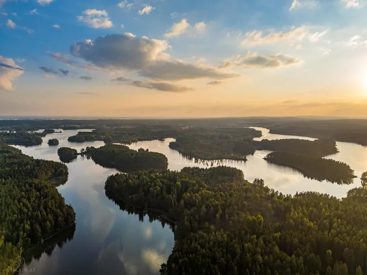 An aerial view of a large body of water surrounded by trees, with the sun setting in the background and clouds in the sky