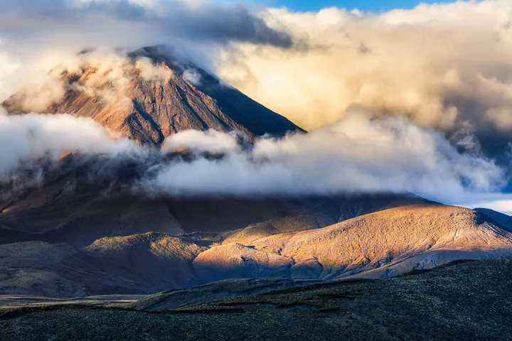 A majestic mountain with a cloud hovering over it, surrounded by lush green grass and a sky filled with white, fluffy clouds