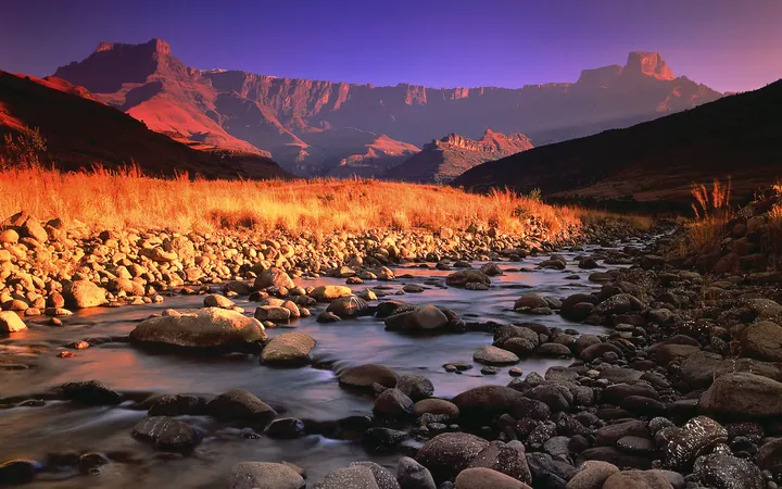 A river running through a rocky valley with mountains in the background, surrounded by lush green grass and a clear blue sky
