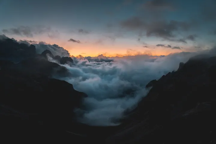 A stunning view of a mountain range covered in clouds at sunset, with the sky in the background illuminated in a beautiful array of oranges, pinks, an