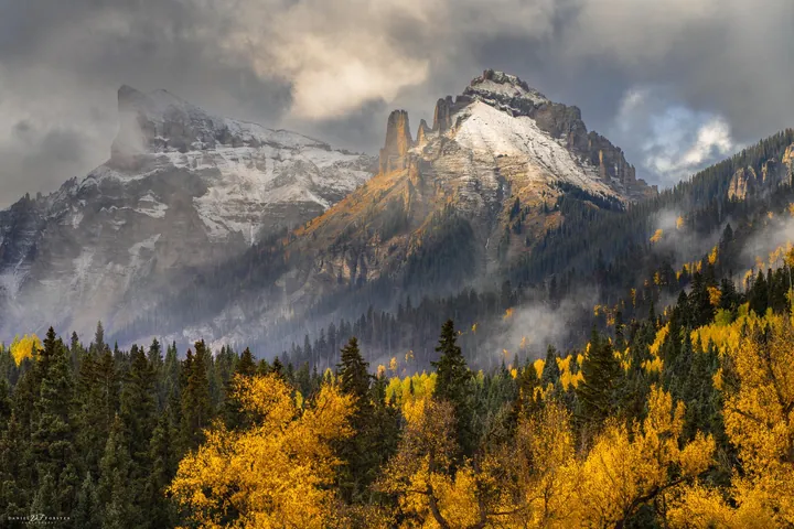 A majestic mountain range with snow-capped peaks in the background, surrounded by trees in the foreground with their leaves changing to vibrant shades