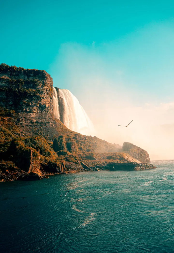 A bird flying over the ocean near a majestic waterfall cascading down a rocky cliff, with a clear blue sky in the background