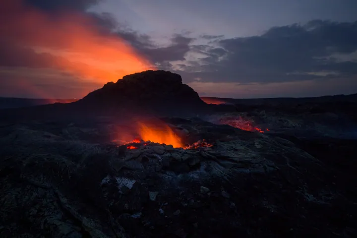 A volcano erupting lava into the ocean at sunset, with a hill in the foreground and a sky filled with clouds in the background