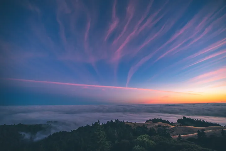 A beautiful sunset over the ocean from the top of a hill, with trees in the foreground and clouds in the sky