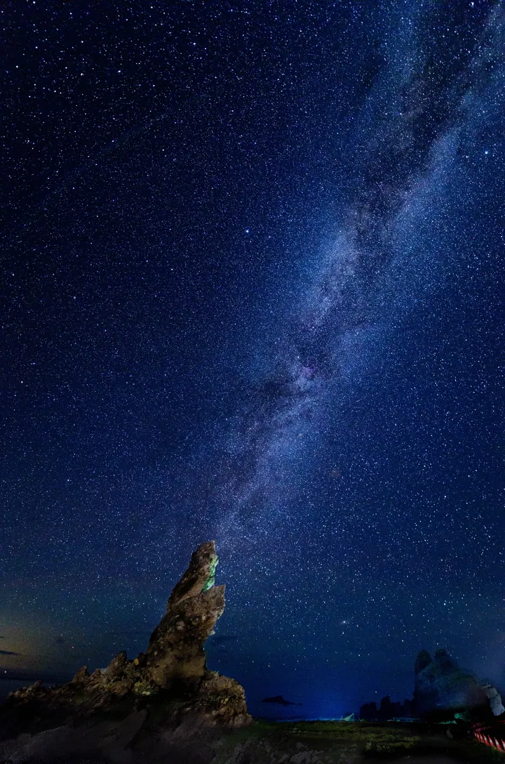 A rock formation in the middle of a field, surrounded by trees and illuminated by the milky way shining brightly in the night sky