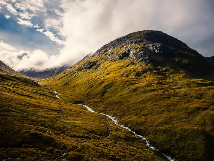 A mountain with a small stream running through it, surrounded by lush green grass and a sky filled with white, fluffy clouds