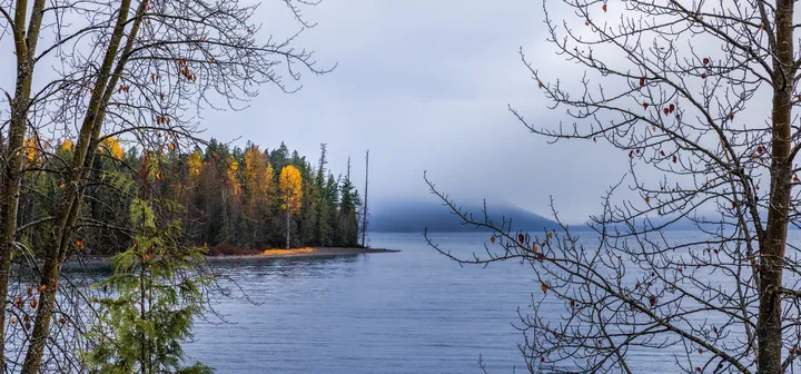 A small island in the middle of a lake surrounded by trees, with a hill in the background and a sky filled with clouds