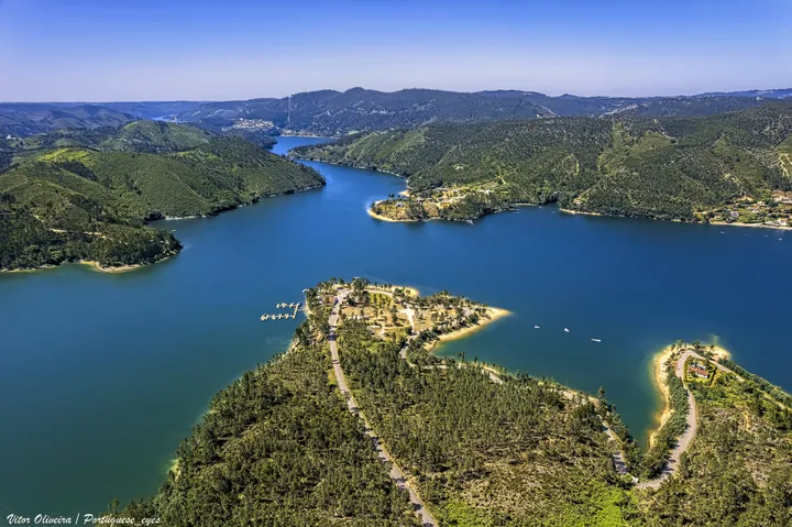 An aerial view of a large body of water surrounded by lush green hills and trees, with a bright blue sky in the background