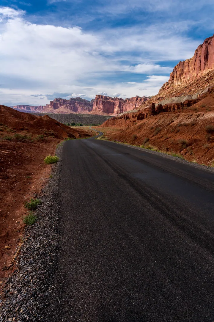 A winding road snaking through a desert landscape, with hills on either side and a sky filled with clouds in the background