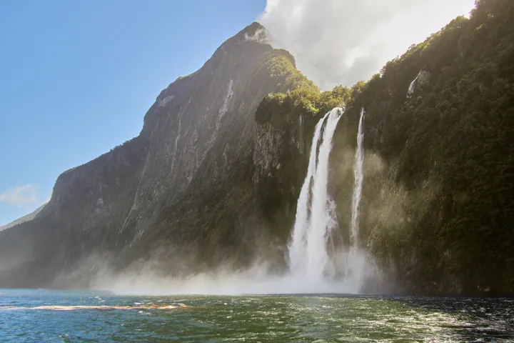Milford Sound in Fiordland National Park, New Zealand, with a majestic waterfall cascading down a hill surrounded by lush green trees and a bright blu