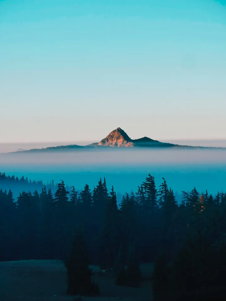 A majestic mountain in the middle of a foggy forest, with trees at the bottom and a beautiful sky in the background