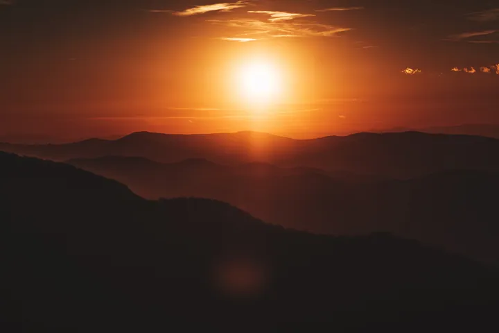 A breathtaking view of the sun setting over the mountains from the top of a mountain, with the sky filled with clouds and the sun casting a warm orang